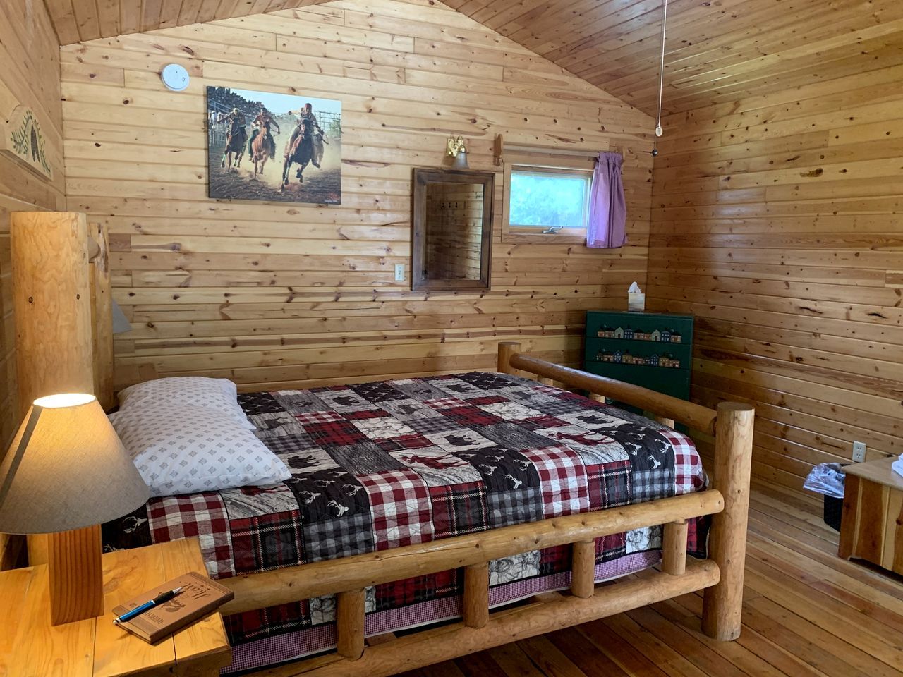 A wooden cabin room with a bed, a nightstand, a lamp, a wall-mounted clock, a framed picture, and a window with curtains.