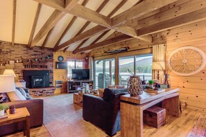 A cozy cabin interior with a fireplace, television, wooden furniture, and a large window offering a mountain view. - Private Dock, Deck & Yard: Luxe Sacheen Lake Home (Newport)