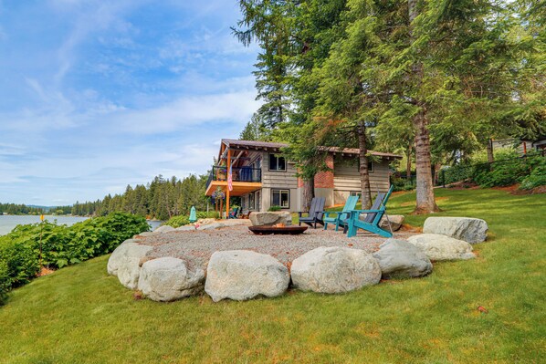 A cabin with a deck, surrounded by large rocks and greenery, overlooking a body of water. - Private Dock, Deck & Yard: Luxe Sacheen Lake Home (Newport)