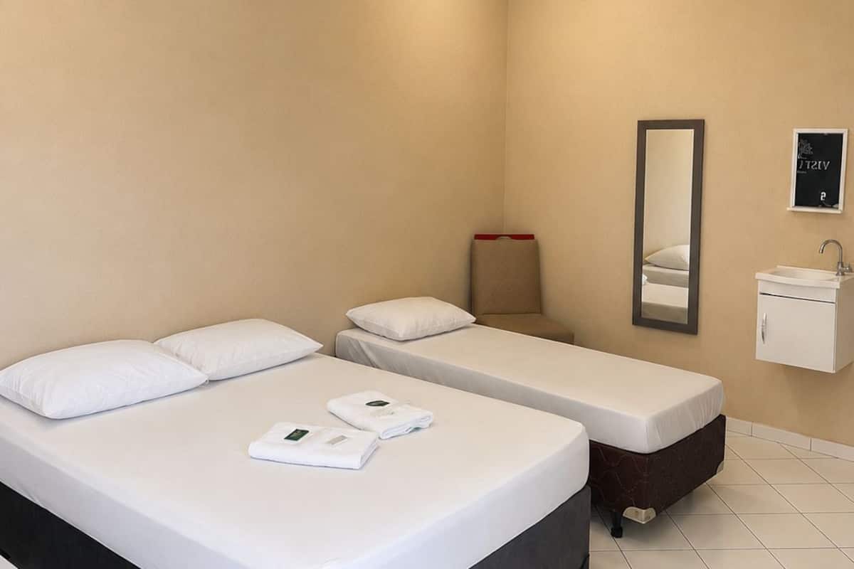 Une chambre d’hôtel avec deux lits simples, une petite table de chevet, un miroir, un lavabo fixé au mur et un tableau encadré.