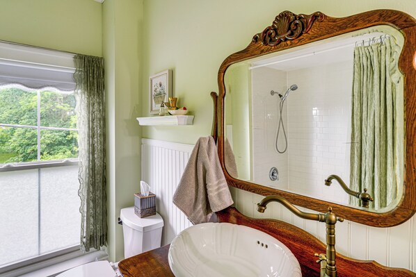 A bathroom with a large mirror, a sink, a shower, and a window with curtains.