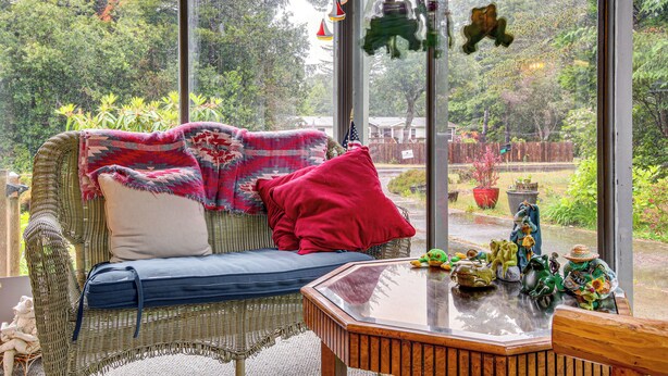 Un sofá de mimbre con una almohada roja y un cojín azul, una mesa de madera con adornos, y vistas de un jardín a través de amplios ventanales.