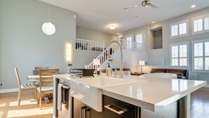 Una cocina moderna con una isla central, mesa de comedor de madera y sillas, una escalera con barandillas blancas y ventanas grandes.