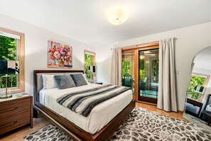 Ein Schlafzimmer mit einem Bett, einem Nachttisch, einer Lampe, einem Gemälde, einem Spiegel und Blick auf einen Garten durch eine Glasdoor.