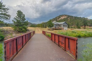 A rustic bridge leading to a cabin with a stone foundation, surrounded by a forested landscape and a river. - Colorado Riverfront Escape! Fish, Hike & Relax (Pine)