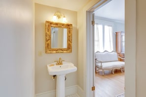 Ein Badezimmer mit einem Standwaschbecken, einem goldfarben gerahmten Spiegel und einer weißen Tür zu einem Raum mit einem weißen Stuhl.