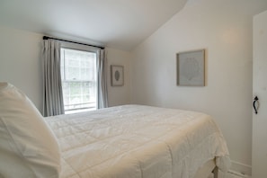 Un dormitorio con cama, ventana con cortinas, un cuadro enmarcado y una lámpara de pared.