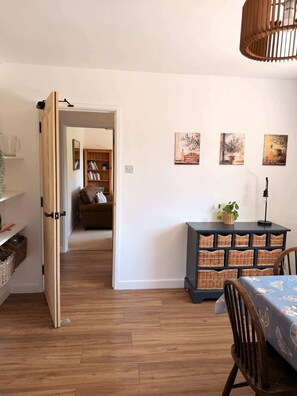 A room with a wooden floor, a dining table with a blue tablecloth, a dark blue cabinet with wicker baskets, and a doorway leading to another room. - Garden Cottage on the edge of Oxford (Kennington)