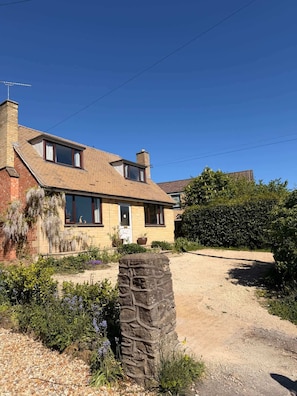 A two-story house with a stone feature in the foreground, a paved driveway, and a garden with various plants. - Garden Cottage on the edge of Oxford (Kennington)