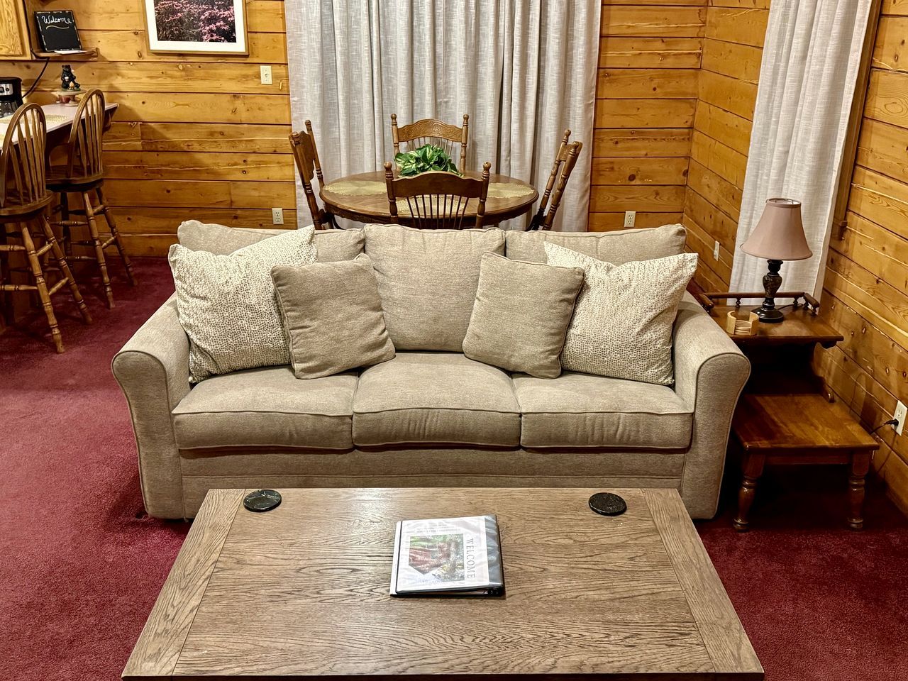 Une pièce aux murs lambrissés, avec un canapé beige, une table basse en bois, une table à manger et des chaises, ainsi qu’une lampe.
