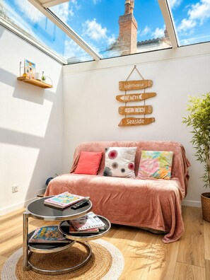 Een gezellige kamer met een roze bank, een klein rond tafeltje met tijdschriften, een houten bord met reisbestemmingen, en een groot raam met uitzicht op een bakstenen schoorsteen.