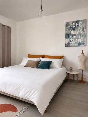 Een netjes opgemaakt bed met wit en bruin beddengoed, een klein nachtkastje met een lamp, en een kunstwerk aan de muur.