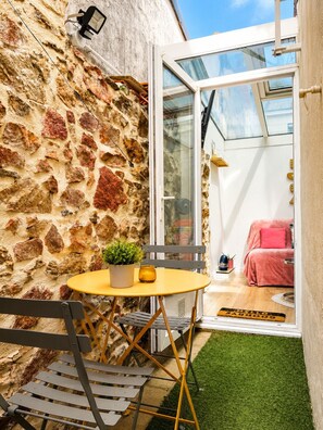 Een klein terras met een stenen muur, een glazen deur die naar een kamer leidt, een ronde tafel met een plant en een bankje.