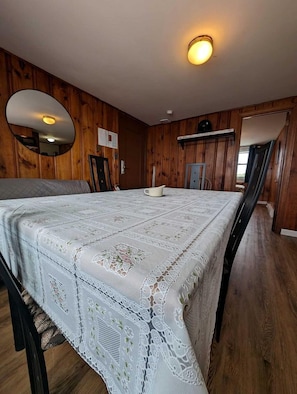 A room with a large dining table covered with a lace tablecloth, wooden walls, a ceiling light, and a wooden floor. - Drift Away in a Peaceful Oceanfront Escape in North Truro, Massachusetts (North Truro)