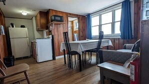 Een kleine keuken met houten kastjes, een witte koelkast en een magnetron. Er is een zithoek met een tafel en stoelen, en een raam met blauwe gordijnen.