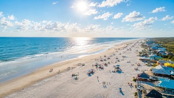 A beach with a row of houses along the shore, a clear sky, and the sun setting over the ocean.