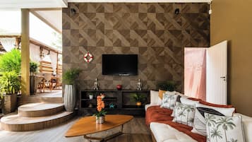 A living room with a patterned wall, a flat-screen TV, a sofa with palm tree pillows, a coffee table, and a staircase leading to an outdoor area.