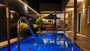 Una piscina con escalera metálica, rodeada por un suelo de baldosas y un área interior con asientos, una banca y una sombrilla.