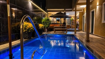 A swimming pool with a metal ladder, surrounded by a tiled deck and indoor seating area with a bench and umbrella.