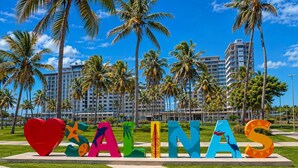 Un letrero con letras de colores que dice 'SA ALNAS' en un parque con palmeras y un hotel al fondo.