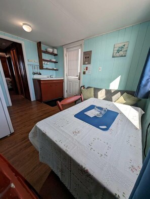 A room with a bed, a small table with a blue placemat, a window with curtains, a door, and a kitchenette area. - Recharge by the Ocean in Total Tranquility in North Truro, Massachusetts (North Truro)
