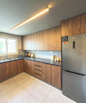 A modern kitchen with wooden cabinets, a stainless steel refrigerator, and a window providing natural light. - Nicosia 2BR Apt with AC & Washer (Metehan)