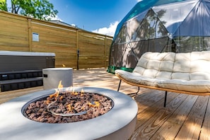 A fire pit with burning wood, a cushioned outdoor chair, and a covered area, all on a wooden deck.