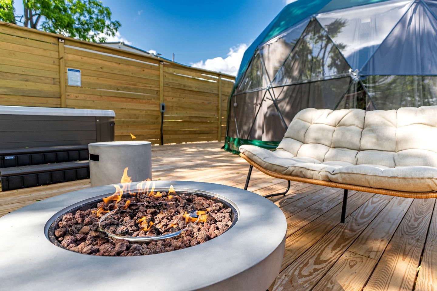 A fire pit with burning wood, a cushioned outdoor chair, and a covered area, all on a wooden deck.