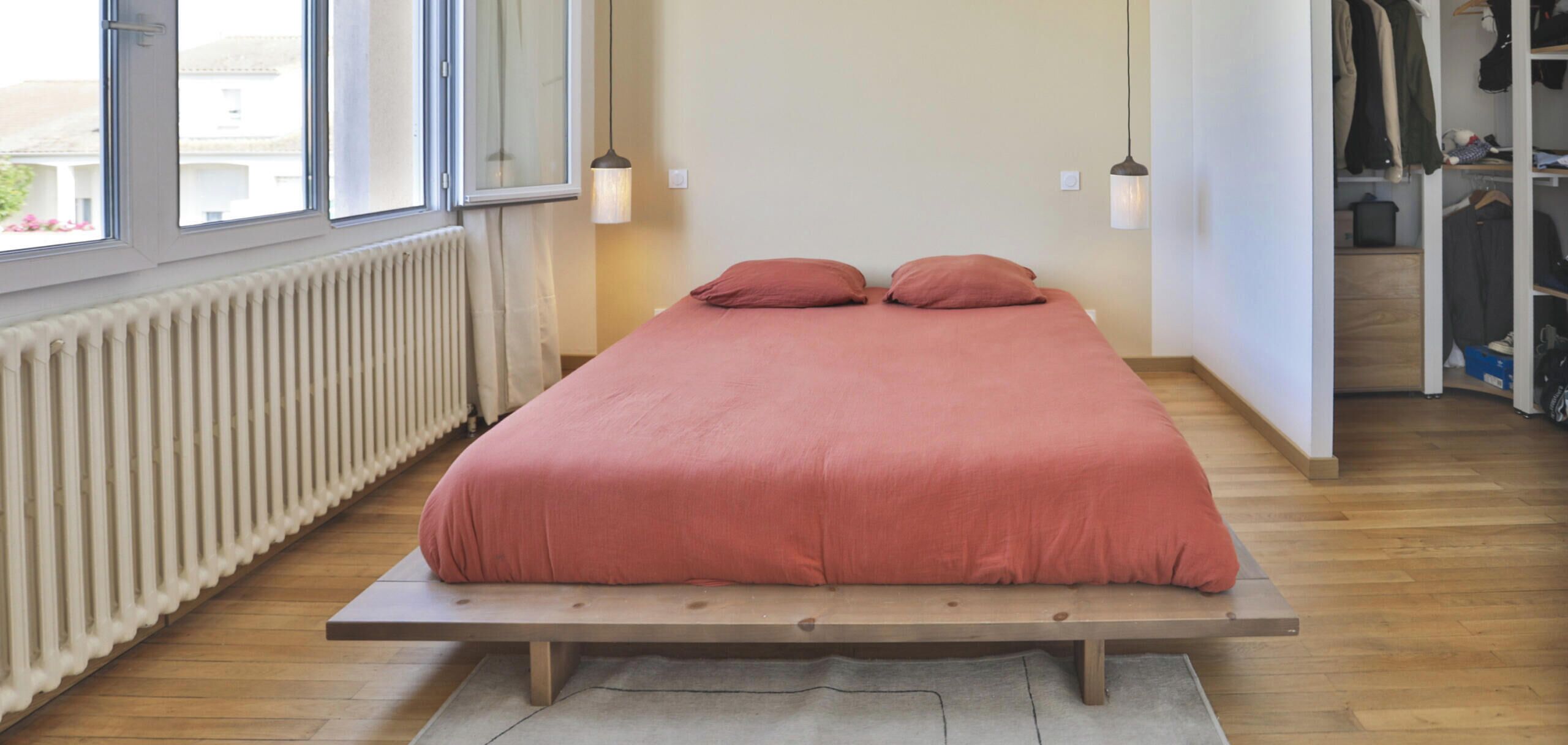 Una cama sobre una plataforma de madera, un radiador, una ventana y un armario con ropa colgada.