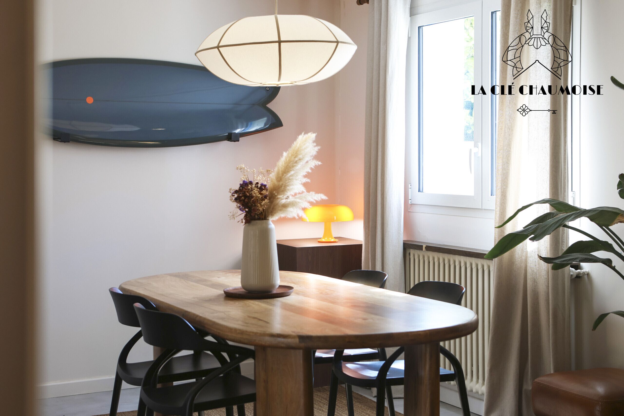 A dining area with a round wooden table, black chairs, a vase with dried flowers, a table lamp, and a large window with curtains.