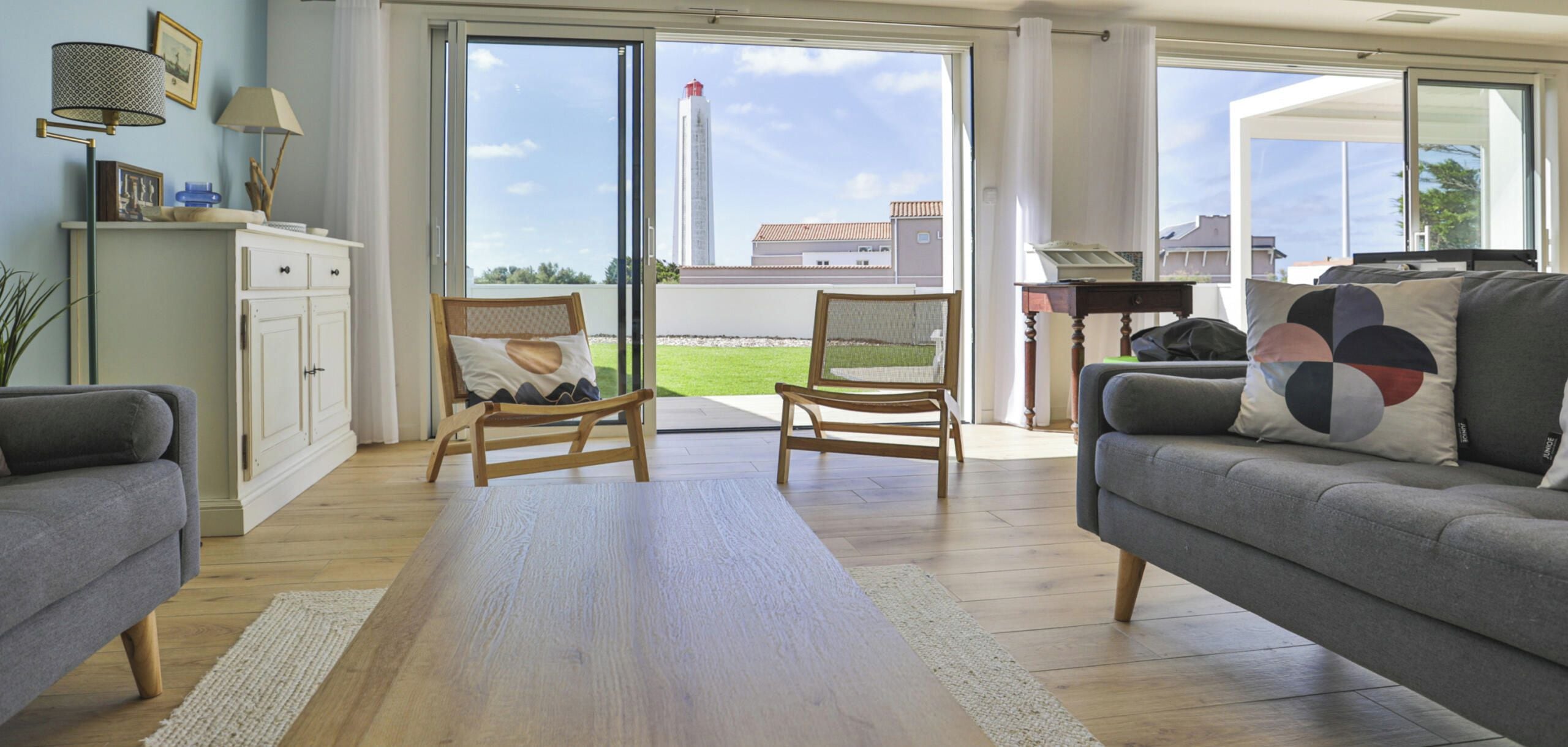Un salon spacieux avec un canapé gris, deux fauteuils en bois, un meuble blanc et une grande fenêtre donnant sur un immeuble et un ciel dégagé.