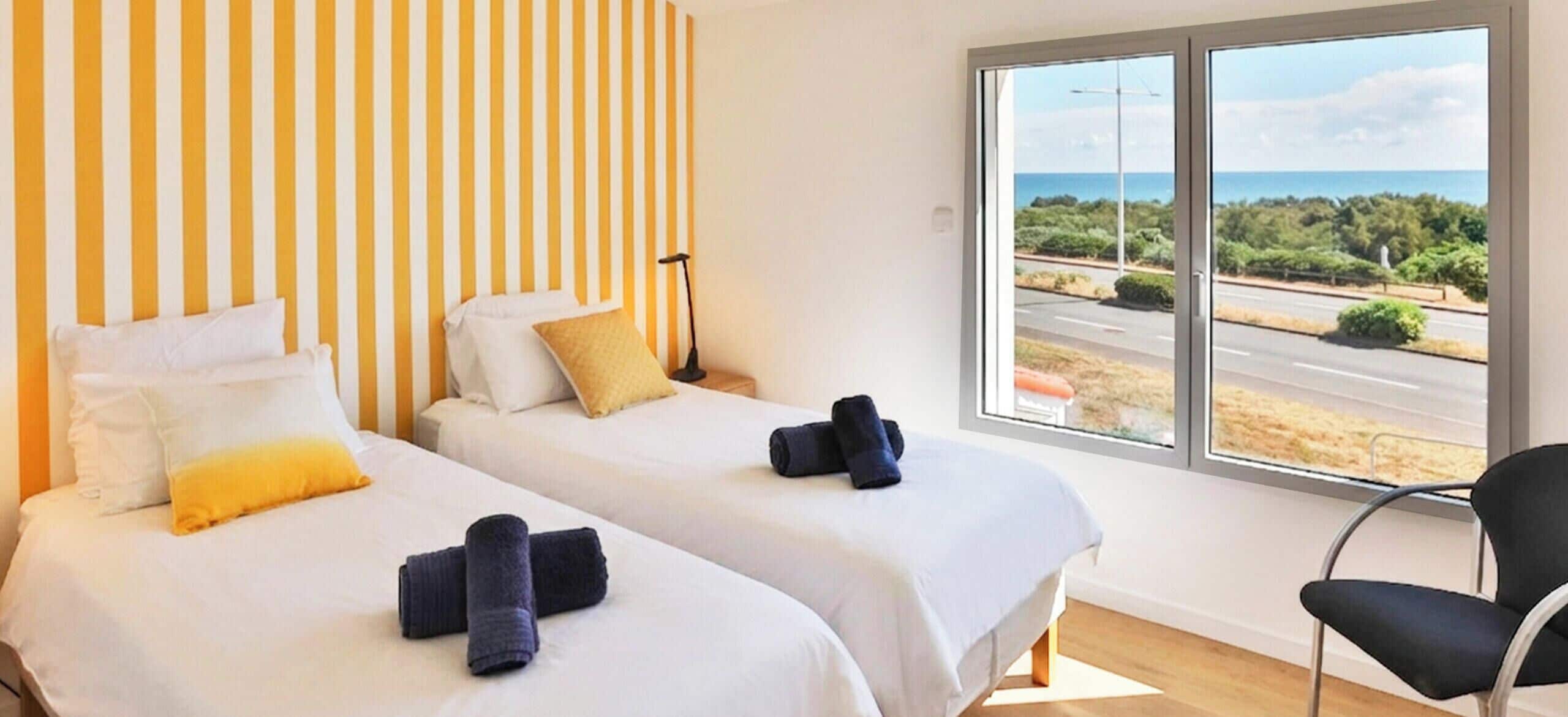 Une chambre d’hôtel avec deux lits, une grande fenêtre donnant sur une route et des espaces verts, et un fauteuil.