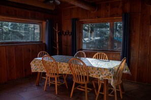 A wooden cabin dining area with a table and chairs, large windows, and a view of the forest. - Cabin #1 (Flagstaff)