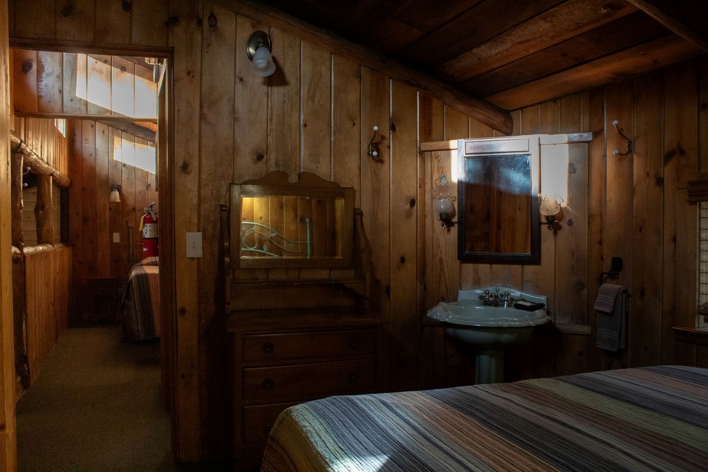 Interior de una cabaña de madera con una cama, un aparador de madera, un espejo y un lavabo.