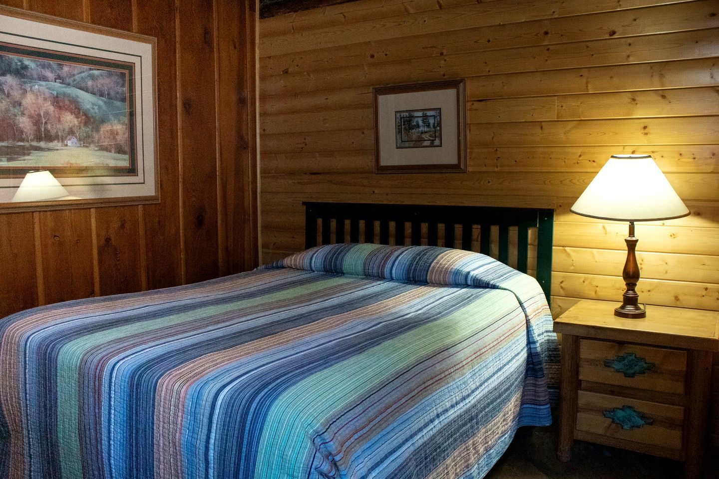 A wooden room with a bed, a nightstand, and two framed pictures on the wall.