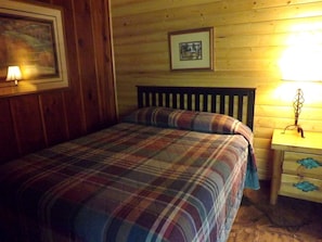 Ein Zimmer mit Holzwandverkleidung, einem Bett, einem Nachttisch, einem gerahmten Bild und einer Wandlampe.