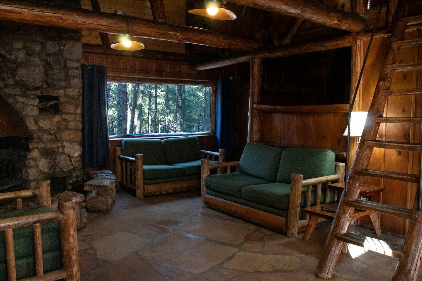 Interior rústico de una cabaña con chimenea de piedra, mobiliario de madera y vistas al bosque a través de una ventana.