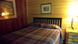 Habitación con paneles de madera, una cama, una mesita de noche, un cuadro enmarcado y una lámpara fijada a la pared.