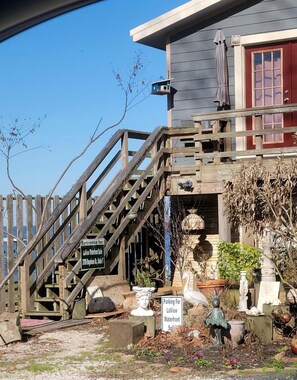 Een houten trap leidt naar een huis met een rode deur, een bordje dat de ingang voor een bepaalde locatie aangeeft, en een standbeeld van een figuur.