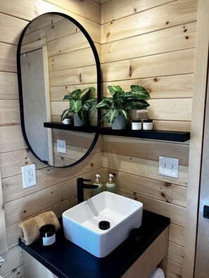 Une salle de bain avec un lavabo blanc, un plan de toilette noir, des murs recouverts de panneaux en bois, un miroir rond et deux étagères ornées de plantes et d’articles de toilette.