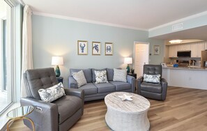Ein Wohnzimmer mit grauem Sofa, zwei Sesseln, einem Couchtisch und einer Küche im Hintergrund.