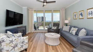 A living room with a flat-screen TV, a ceiling fan, a sofa, a coffee table, and a view of the city through large windows. - New to Program - Silver Shells St. Lucia 602 (Destin)