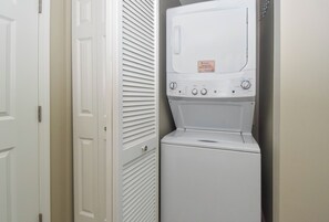 A compact, white laundry room with a front-loading washer and dryer, a door with a handle, and a wall-mounted vent. - Lagoon Pool- The Palms of Destin 2303 by Compass Resorts (Destin)