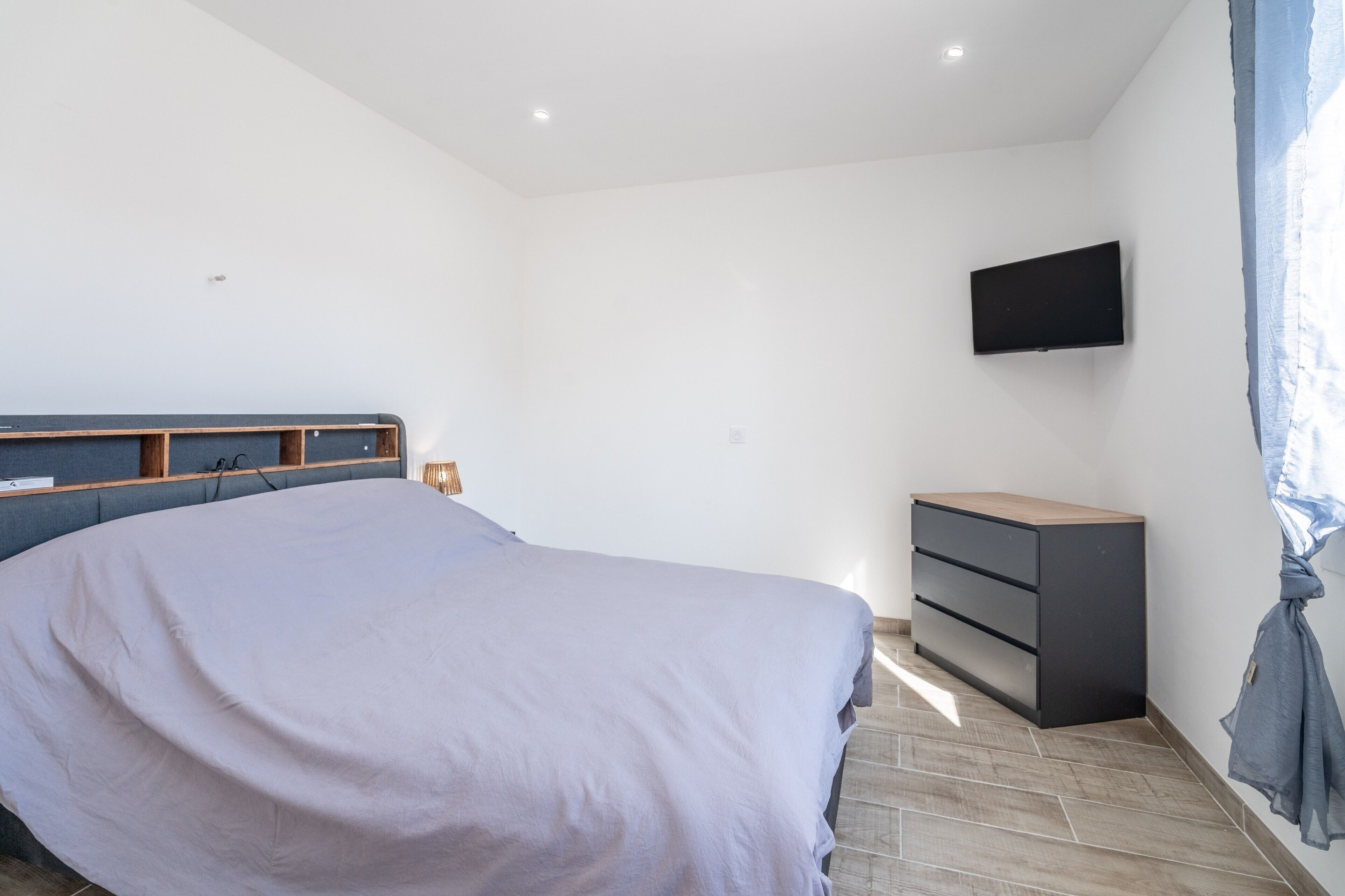 A bedroom with a bed, a TV mounted on the wall, a dresser, and a window with curtains.
