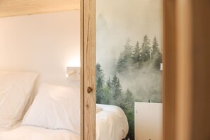 Ein Schlafzimmer mit einem Bett, einem Nachttisch und Blick auf einen Wald durch ein Fenster.