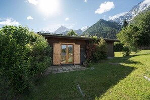 Une cabane en bois avec une porte en verre, entourée d’une végétation luxuriante et avec des montagnes en arrière-plan.