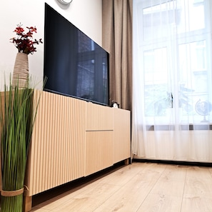 Ein modernes Wohnzimmer mit Fernseher an der Wand, einem Holzschrank, einer Zimmerpflanze und einem großen Fenster mit Vorhängen.