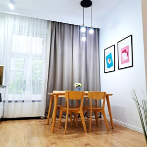 Ein Essbereich mit Holztisch und Stühlen, einem Fenster mit durchsichtigen Vorhängen, einem grauen Vorhang und zwei gerahmten Bildern an der Wand.