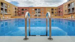 Una piscina con escaleras de metal, rodeada por un deck de baldosas, y un edificio de varios pisos con balcones al fondo.