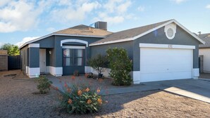 A single-story house with a garage, a small front yard with a gravel driveway, and a clear sky. - Group Home Near Stadium | Sleeps 14+ (El Mirage)